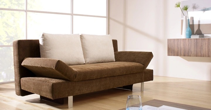 Galerie-Bild 5: Braunes Sofa mit beigen Kissen steht vor großen Fenstern in einem modernen Wohnzimmer. Im Vordergrund steht ein Tisch mit Glas Wasser und einem geöffneten Buch, Vasen sind im Hintergrund.
