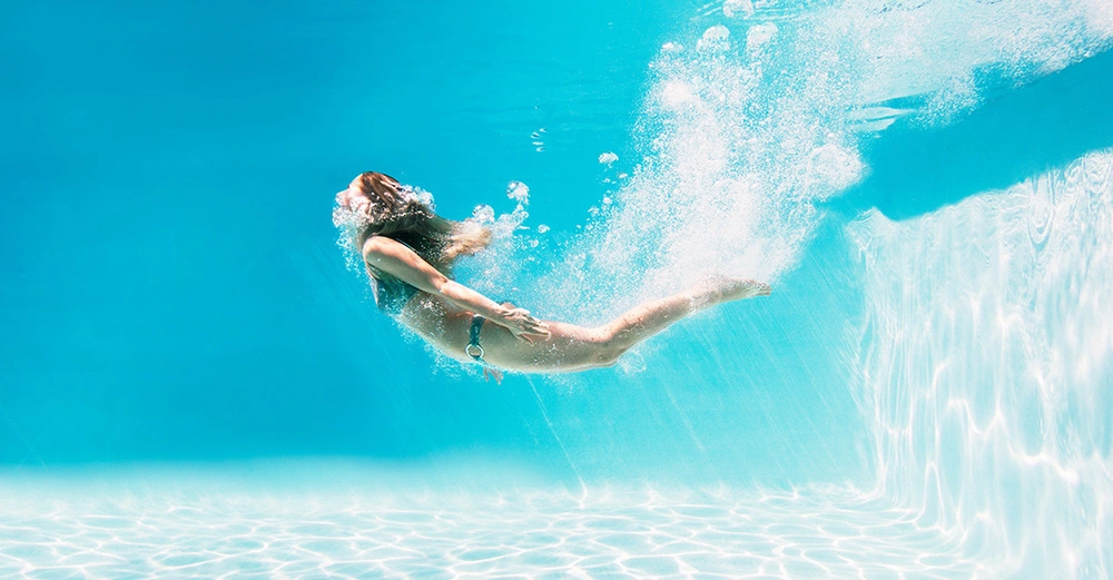 Frau taucht unter Wasser in einem klaren Pool. Sommerliche Szene mit spritzendem Wasser und Sonnenlicht.