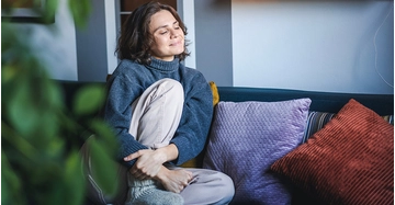 Eine Frau mit braunen Haaren und blauem Pullover sitzt auf einer gemütlichen Couch in ihrem Wohnzimmer und genießt die gesunde Wärme ihrer Infrarotheizung.