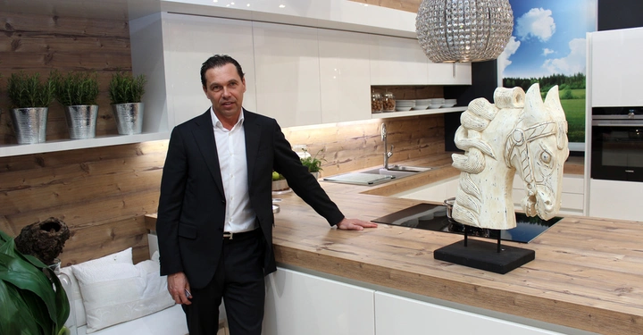 Galerie-Bild 25: Ein Mann steht in einer modernen Küche neben einer Arbeitsplatte und einer Pferdeskulptur; die Küche hat Holzoberflächen, weiße Schränke, Pflanzen und eine große Hängeleuchte.