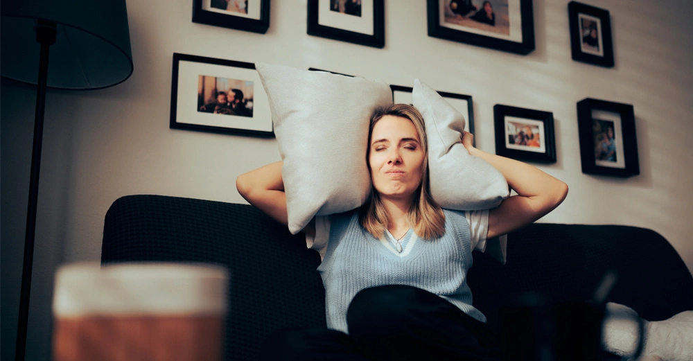 Genervte Frau mit zugekniffenen Augen sitzt auf dem Sofa und presst sich zwei weiße Kissen an die Ohren.