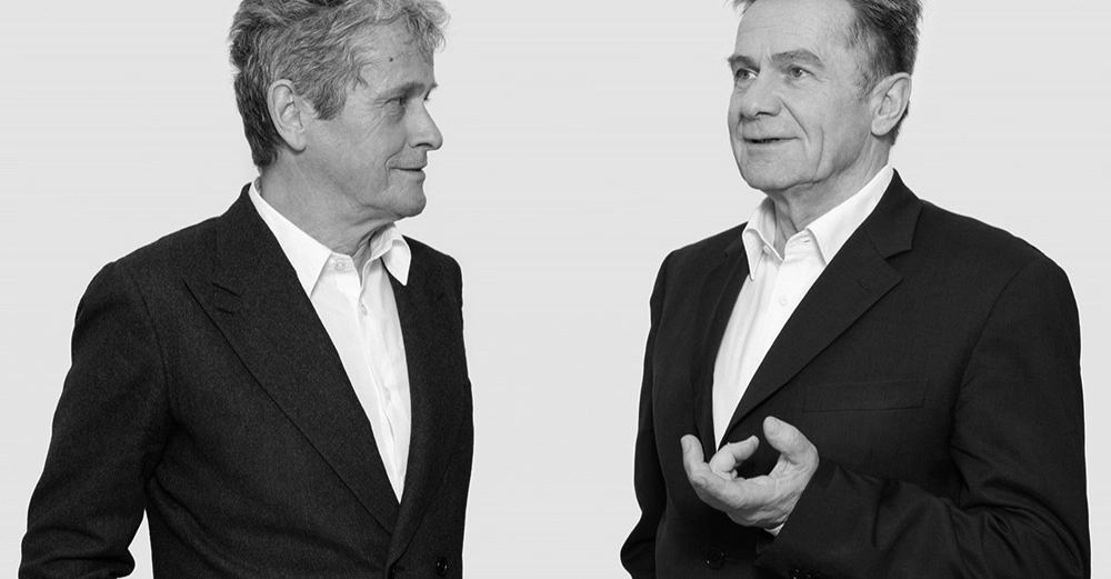 Zwei Männer in Anzügen unterhalten sich stehend vor einem hellen Hintergrund. Der Mann links schaut lächelnd auf den Mann rechts, der sprechend eine Hand bewegt.