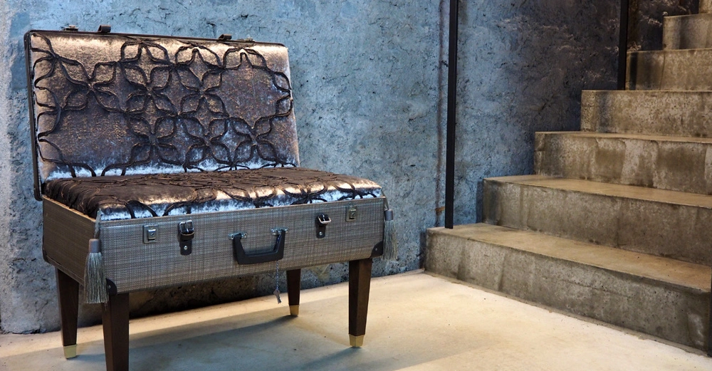 Alter Koffer dient als Stuhl im Betonraum neben Betontreppe, mit grauem Kissen und floralem Muster auf der Rückseite