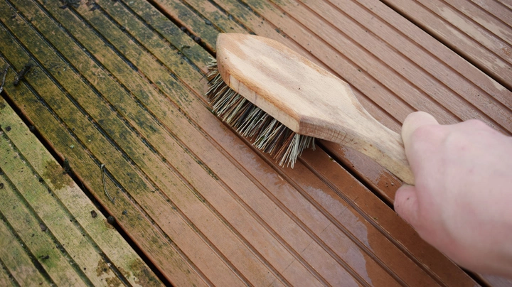 Eine Hand reinigt mit einer Bürste verwitterte und teils vermooste Holzdielen einer Terrasse. Der gereinigte Bereich wirkt deutlich heller und gepflegter.