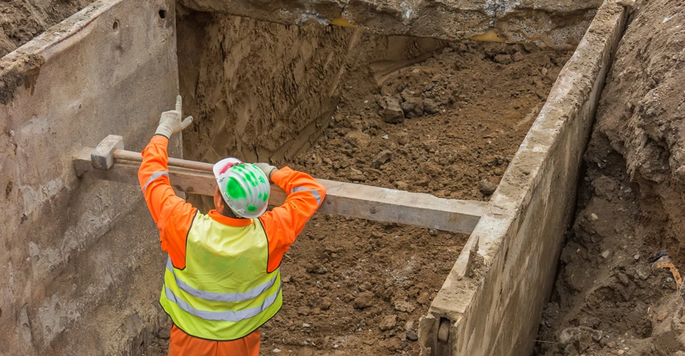 Bauarbeiter in orangefarbener Schutzkleidung richtet eine Holzstrebe zwischen zwei Betonwänden in einem tiefen Erdgraben aus umgeben von Erde und Bauschutt