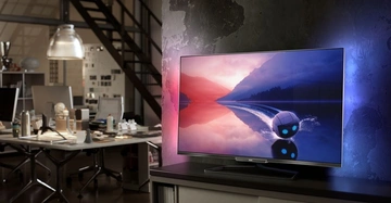 Ein Flachbildfernseher zeigt eine animierte Szene auf einem Schreibtisch in einem Arbeitszimmer mit Regalen, Büchern und einem offenen Loft-Design im Hintergrund.