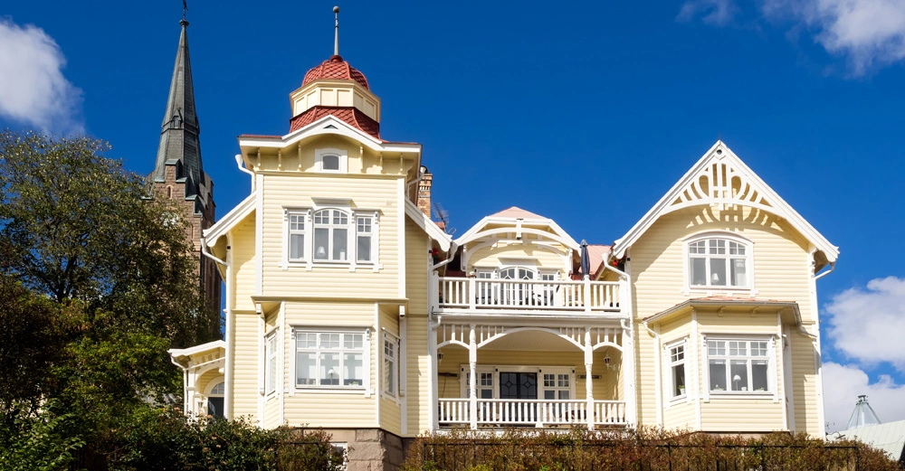Hellgelbes historisches Haus steht ruhig vor klarem blauen Himmel. Im Hintergrund ragt der Turm eines hohen Gebäudes hervor, umgeben von Bäumen und Büschen.