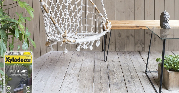 Eine weiße Hängematte hängt neben einem Holztisch mit Glasplatte in einem Raum mit rohem Holzboden; links steht eine Pflanze neben einer Xyladecor-Dose mit dem Text Xyladecor Garten GARDEN FLÄIRS. Imprägniertes Holzöl für eine schützende natürliche Optik.