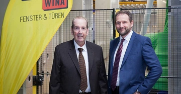 Zwei Männer in Anzügen stehen nebeneinander, lächelnd in einer modernen Fabrikumgebung. Links ein gelbes Schild mit der Aufschrift WINA FENSTER & TÜREN. Im Hintergrund Maschinerie und Metallgitter.