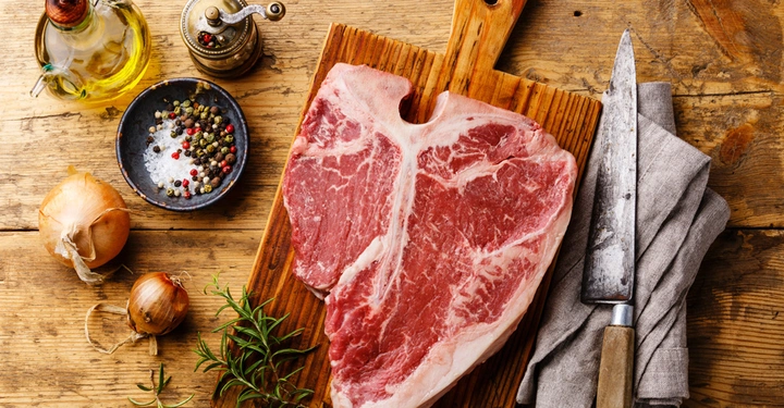 Ein großes rohes Steak liegt auf einem hölzernen Schneidebrett Küchenmesser nebenan umgeben von einer Ölkaraffe einer Pfeffermühle Zwiebeln Rosmarin und einer Schale mit Salz und Pfefferkörnern auf einem hölzernen Tisch