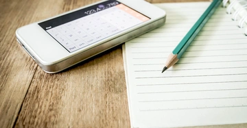 Smartphone zeigt Taschenrechner liegend auf Holztisch daneben ein Notizbuch mit grünem Bleistift liegend auf Linienpapier in einem hellen Raum.