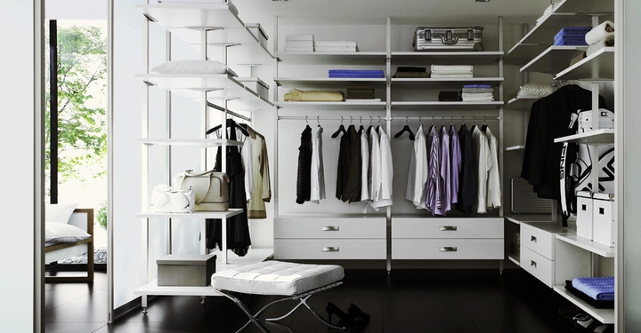 Galerie-Bild 8: Ein begehbarer Kleiderschrank mit weißen Regalen und Schubladen organisiert Kleidungsstücke und Accessoires. Ein weiße Bank steht inmitten, während helle Tageslicht durch große Fenster strömt.