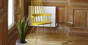 Gelber Stuhl steht neben einem kleinen runden Tisch mit goldenem Tablett in einem Raum mit Holzwänden und -boden; links von ihm steht eine grüne Pflanze in einem weißen Topf.