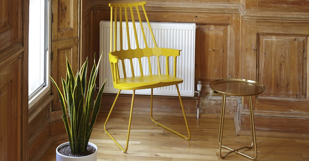 Gelber Stuhl steht neben einem kleinen runden Tisch mit goldenem Tablett in einem Raum mit Holzwänden und -boden; links von ihm steht eine grüne Pflanze in einem weißen Topf.