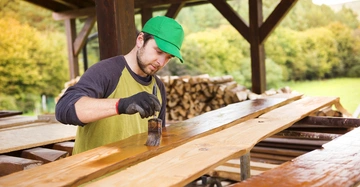 Mann mit grünem Hut streicht Holzplanke mit Pinsel in überdachter Freiluftwerkstatt umgeben von Bäumen und Holzstapeln.