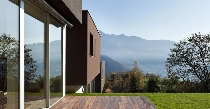Modernes Haus mit großen Glasfenstern und Holzbalkon steht in einer grünen Landschaft mit Bäumen vor einem Panorama aus Bergketten und unter wolkenlosem Himmel.