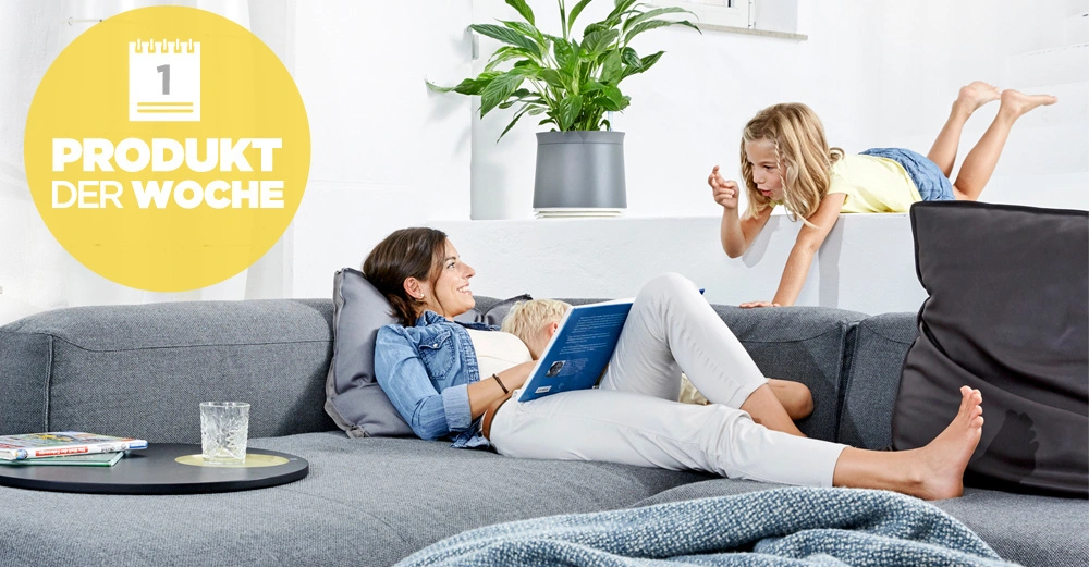 Frau liest ein Buch auf einem grauen Sofa. Kind klettert auf die Rückenlehne. Heller Raum mit Pflanze im Hintergrund. Text im gelben Kreis: 1 PRODUKT DER WOCHE.