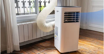 Eine mobile Klimaanlage in einer Altbauwohnung, die kühle Luft im Raum verteilt und gleichzeitig die heiße Luft durch einen Abluftschlauch durch das vergitterte Fenster nach draußen leitet.