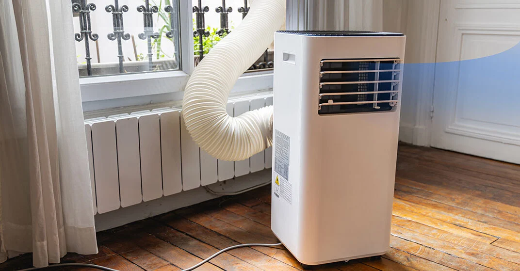 Eine mobile Klimaanlage in einer Altbauwohnung, die kühle Luft im Raum verteilt und gleichzeitig die heiße Luft durch einen Abluftschlauch durch das vergitterte Fenster nach draußen leitet.