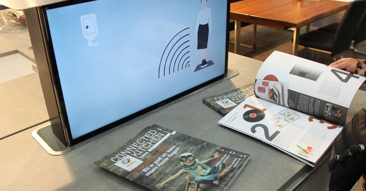 Galerie-Bild 20: Monitor zeigt eine Person und ein Signal in einer Messeumgebung auf einem grauen Tisch mit Broschüren CONNECTED KITCHEN und einer lesenden Person. Text auf Broschüre: VISIEREN UND ERLEBEN Bis es "good to scan" unsere Küche finden