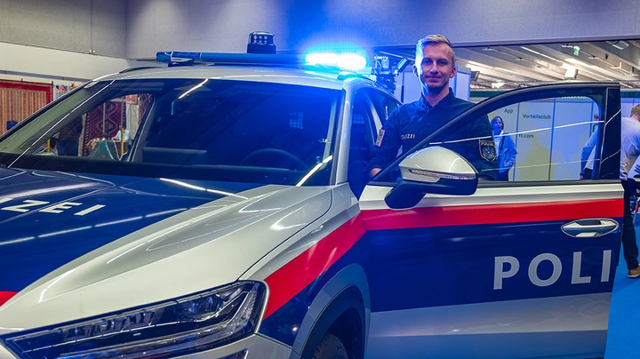 Galerie-Bild 3: Polizist steht hinter geöffneter Wagentür vom Polizeiauto und lächelt in die Kamera.
