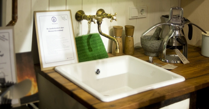 Galerie-Bild 5: Ein Waschbecken mit messingfarbenem Wasserhahn steht auf einer Holzarbeitsplatte in einer Küche. Daneben sind eine grüne Reinigungsbürste Holzmühlen ein Mörser und eine Saftpresse. Text: So funktioniert der Wasserkreislauf