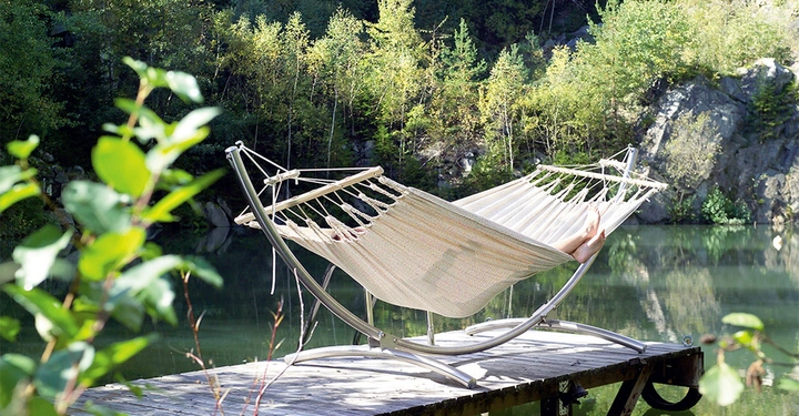 Galerie-Bild 2: Hängematte ruht auf Gestell Füße ragen heraus Hütte liegt am ruhigen See umgeben von Bäumen und Felsen