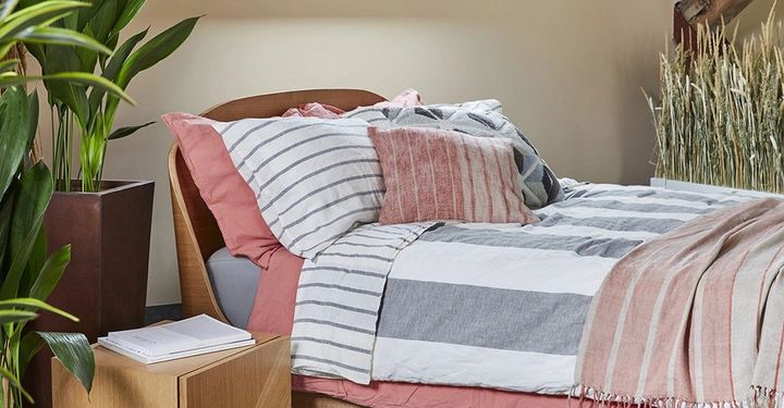 Bett mit gestreifter Bettwäsche und Kissen steht in einem Schlafzimmer neben einem Nachttisch mit Büchern und einer großen Pflanze in einem Blumentopf im Hintergrund befinden sich Dekorationsgräser