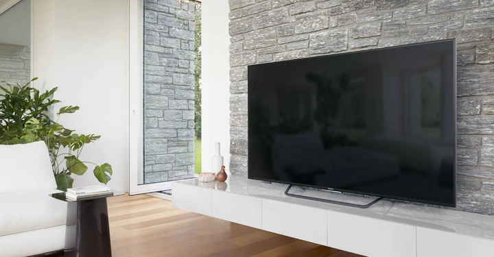 Galerie-Bild 3: Flachbildfernseher stehend auf weißem modernen Schrank vor einer Steinwand in einem hellen, minimalistischen Wohnzimmer mit Holzboden, einer Pflanze im Topf, einem weißen Sessel und einem kleinen schwarzen Beistelltisch mit Büchern