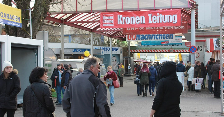 Galerie-Bild 4: Menschen gehen unter einem überdachten Gang bei einer Messe viele Banner und Schilder aufgehängt. Text Kronen Zeitung ORF OO MEIN LAND MEIN RADIO OONachrichten www.laumer.at Lindpointner Garagen.