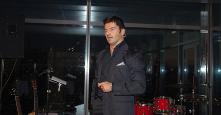 Galerie-Bild 68: Mann im Anzug spricht und benutzt ein Mikrofon stehend auf einer Bühne in einem modernen Raum mit Musikinstrumenten wie Gitarren und einem Schlagzeug im Hintergrund