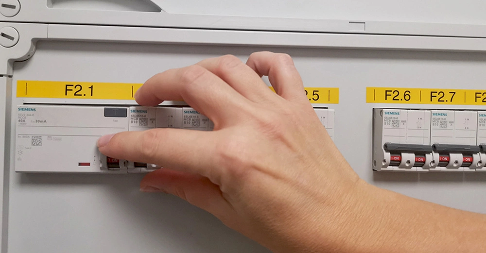 Hand justiert einen Siemens-Sicherungsschalter in einem Schaltschrank. Umgeben von gelben Labels F2.1 bis F2.7.