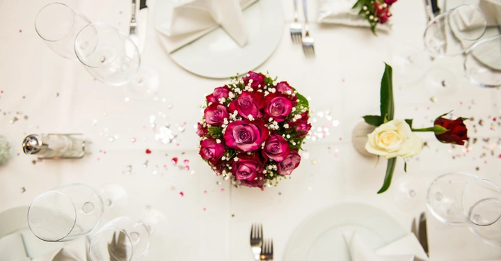 Rote Rosen in einer Vase stehen auf einem festlich gedeckten Tisch mit weißen Tellern Glaswaren und Besteck umgeben von Streudeko und weißen Servietten.