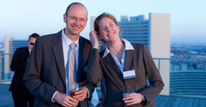 Galerie-Bild 16: Zwei Personen in Anzügen stehen lächelnd auf einem Dach und halten Getränke in der Hand; im Hintergrund sind weitere Personen und moderne Gebäude zu sehen