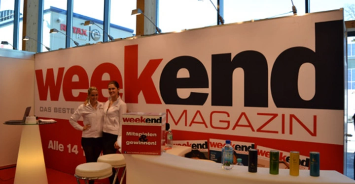 Galerie-Bild 37: Zwei Frauen stehen lächelnd an einem Stand mit Wochenendmagazin-Logo im Hintergrund in einer Messehalle mit hohen Fenstern. Text: weekend MAGAZIN DAS BESTE NUR ALLE 14 Tage Mitspielen und gewinnen.