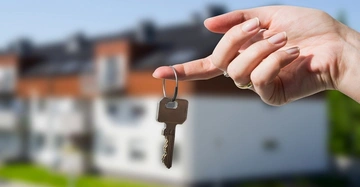 Frauenhand hält Haustürschlüssel für neues Haus, im Hintergrund eine Siedlung mit Reihenhäusern