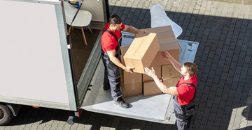 Zwei Männer heben Pappkartons aus einem LKW auf eine Hebebühne auf einem gepflasterten Hof.