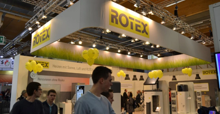 Galerie-Bild 16: Mehrere Menschen gehen an einem Ausstellungstand von ROTEX vorbei Um den Stand herum hängen gelbe Luftballons Text auf dem Banner Heizen mit Sonne Luft und ROTEX Renovieren ohne Risiko