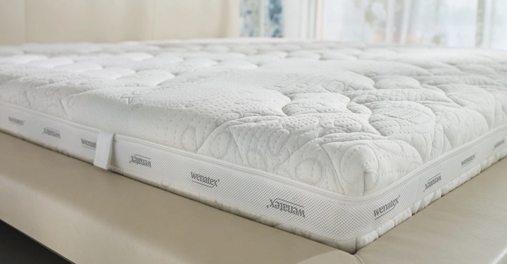 Eine weiße Matratze liegt auf einem beigen Bett. Die Matratze hat ein strukturiertes Muster und die Aufschrift "Wenatex" an den Seiten. Im Hintergrund sind Fenster mit leichter Bepflanzung.