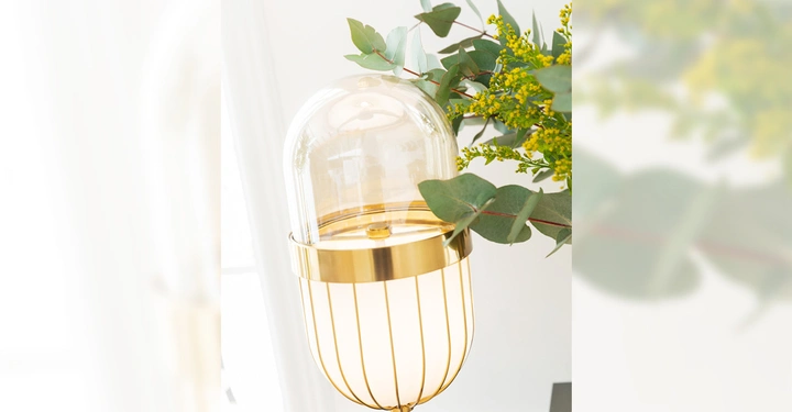 Galerie-Bild 8: Goldene Lampe strahlt sanftes Licht aus, umgeben von grünen Eukalyptusblättern und gelben Blüten, in einem hellen Raum mit weißen Wänden