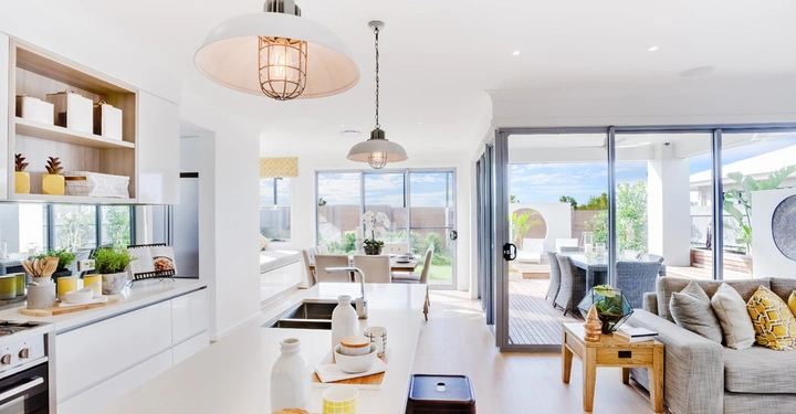 Helle Küche mit Insel und Arbeitsplatte voll mit Geschirr und Pflanzen. Moderne Hängeleuchten. Großer Wohnbereich im Hintergrund mit Sofa und Essbereich. Glas-Terrassentüren führen nach draußen.