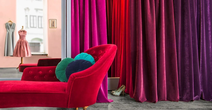 Galerie-Bild 1: Eine rote Chaiselongue mit bunten Kissen steht vor violetten Vorhängen in einem Raum Ein Schaufenster im Hintergrund zeigt zwei Kleider auf Schneiderbüsten und ein Paar Schuhe ist am Boden sichtbar