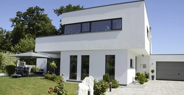 Ein zweistöckiges modernes Haus mit weißen Wänden steht im Zentrum, umgeben von gepflegtem Rasen, Bäumen und einem Steinweg; eine Terrasse mit Sonnenschirm und Liegestühlen befindet sich links.