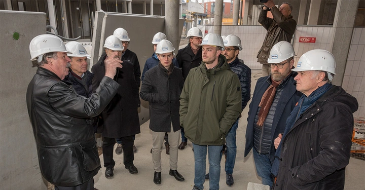 Galerie-Bild 2: Eine Gruppe von Personen mit weißen Bauhelmen steht aufmerksam um einen Mann herum der etwas erklärt in einem unfertigen Gebäude mit Betonwänden und Bauausrüstung.