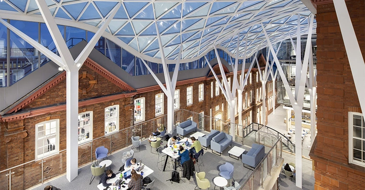 Galerie-Bild 2: Menschen arbeiten in einem modernisierten Gebäude mit Glasdach und roten Backsteinwänden. Innerhalb der Glasstruktur befinden sich Tische, Stühle und Sofas, die eine moderne Arbeitsumgebung schaffen