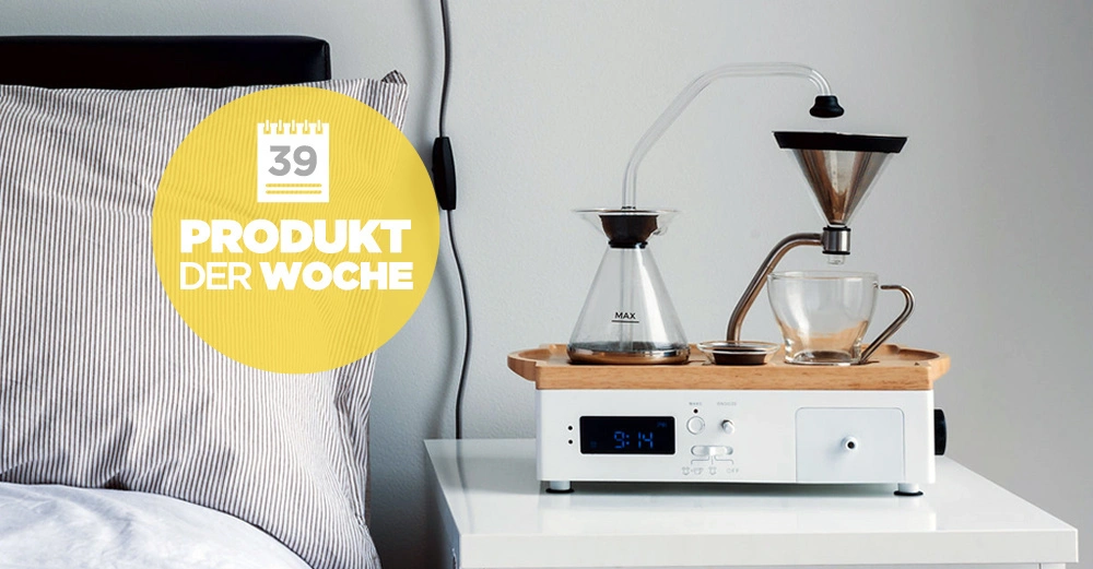 Kaffeemaschine brüht Kaffee auf einem Nachttisch neben einem Bett 39 PRODUKT DER WOCHE in gelbem Kreis Text auf Kaffeemaschine zeigt Uhrzeit 9:14