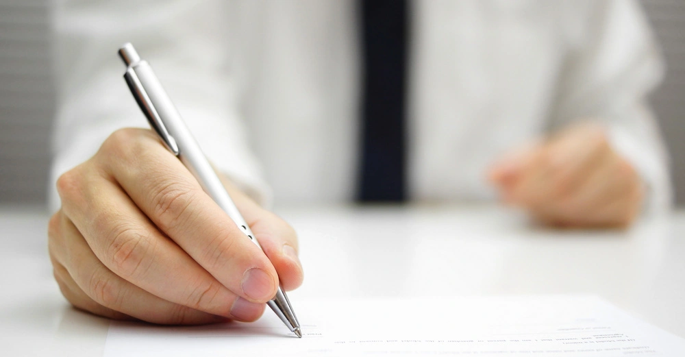 Hand schreibt mit einem Stift auf Papier Person trägt ein weißes Hemd mit Krawatte Hintergrund unscharf Büroumgebung ohne Text.