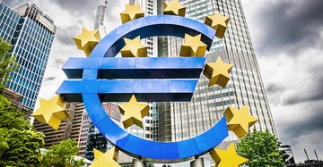 Große blaue Euro-Skulptur mit gelben Sternen steht vor modernen Hochhäusern umgeben von Bäumen unter bewölktem Himmel