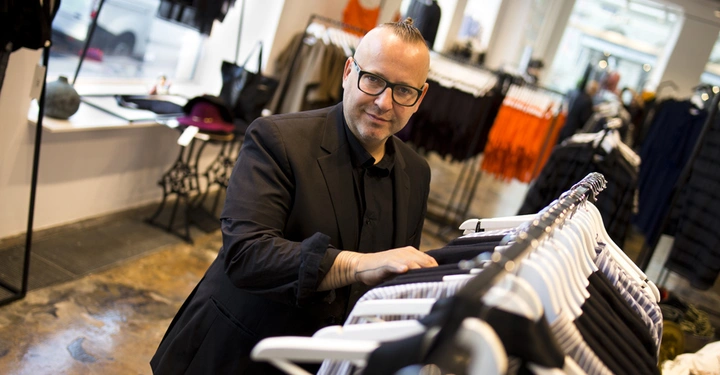 Galerie-Bild 1: Ein Mann in schwarzer Kleidung durchsucht Kleidungsstücke an einem Kleiderständer in einem modernen Bekleidungsgeschäft mit großen Fenstern und unterschiedlichen Kleidungsstücken um ihn herum.