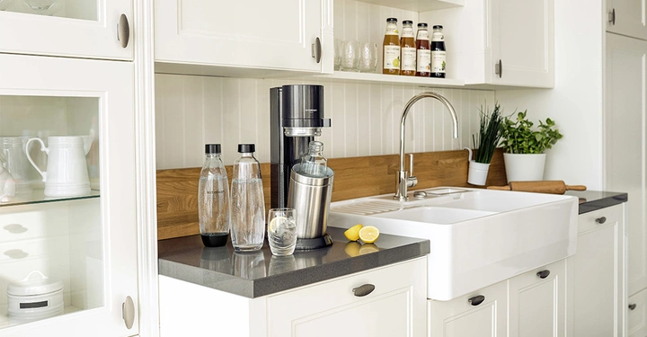 Sodastream-Gerät sprudelt Wasserflasche in heller, moderner Küche mit weißem Schrank, schwarzer Arbeitsplatte, Spüle, Pflanzen, Gläsern, Sirupflaschen und halbiertem Zitrone auf der Arbeitsplatte.